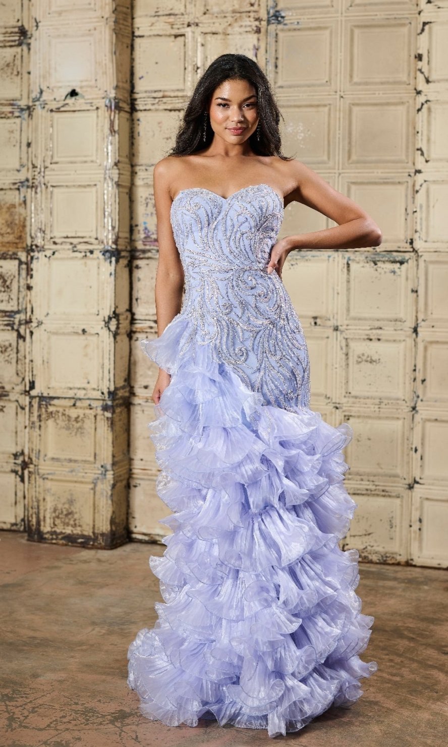 Indoors, a woman wears the Tiffany 16234 Long Prom Dress in lavender, featuring a strapless beaded bodice and layered tulle skirt with intricate detailing and ruffled cascades.